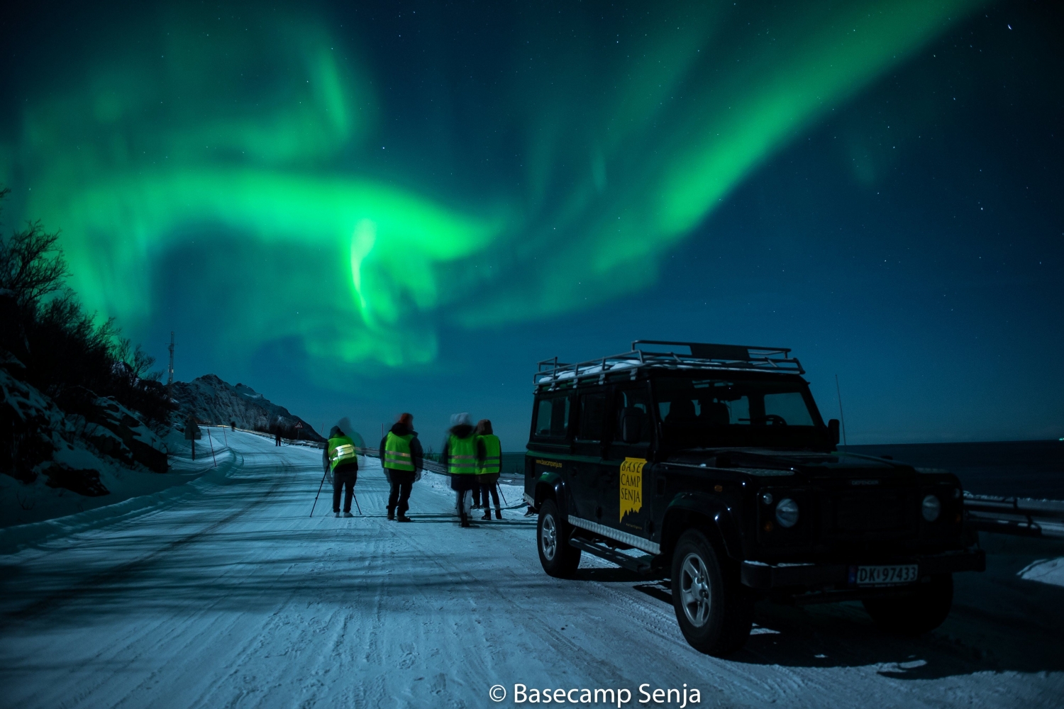 A truck parked on a snowy road with people and Northern Lights in the background