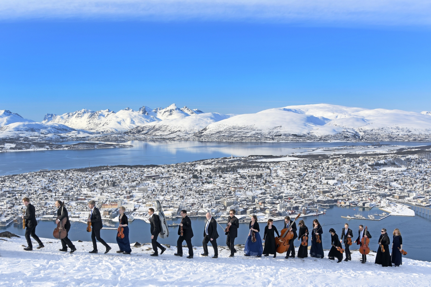 Et tog av musikere som bører deres instrumenter på toppen av Fløya-fjellet, utsikt over Tromsøya i bakgrunnen