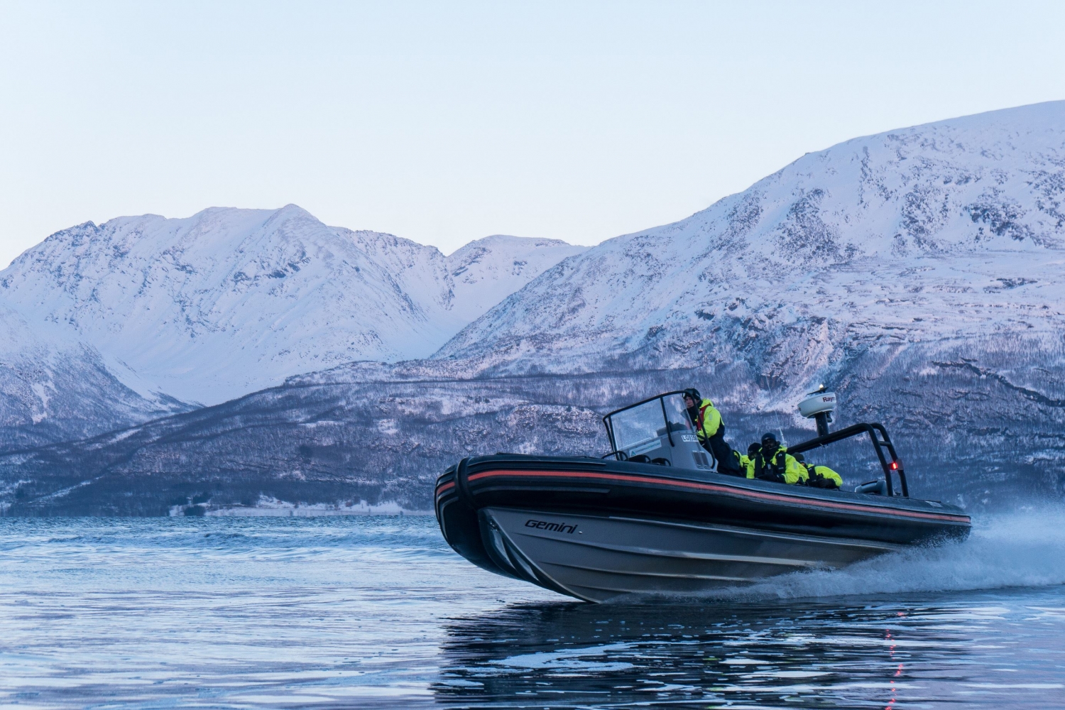 En RIB-båt kjører fort i fjorden med snødekte fjell i bakgrunnen