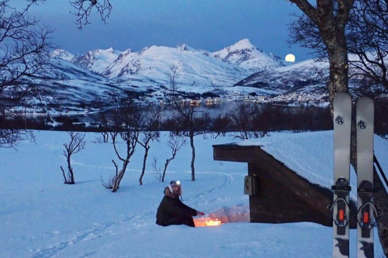 Person nyter bål i snøen mens månen er bak fjellene